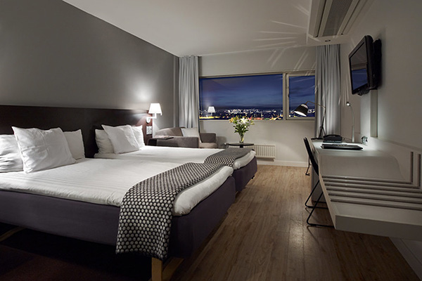 A modern hotel room with two neatly made twin beds, a wall-mounted TV, a desk with a chair, and a large window overlooking a city at night. The room has wooden floors, gray and white decor, and soft lighting from a bedside lamp.