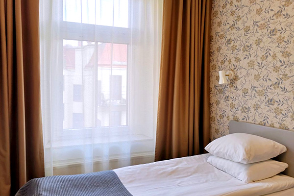 A modern hotel room with two neatly made twin beds, a wall-mounted TV, a desk with a chair, and a large window overlooking a city at night. The room has wooden floors, gray and white decor, and soft lighting from a bedside lamp.