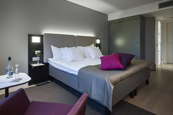 Gothia Towers Standard 2 A modern hotel room with two neatly made twin beds, a wall-mounted TV, a desk with a chair, and a large window overlooking a city at night. The room has wooden floors, gray and white decor, and soft lighting from a bedside lamp.