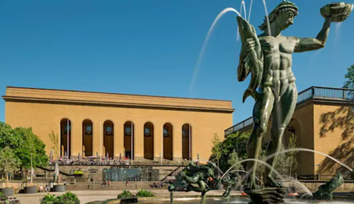 A modern hotel room with two neatly made twin beds, a wall-mounted TV, a desk with a chair, and a large window overlooking a city at night. The room has wooden floors, gray and white decor, and soft lighting from a bedside lamp.