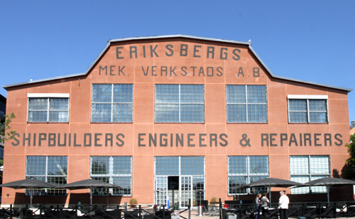 A modern hotel room with two neatly made twin beds, a wall-mounted TV, a desk with a chair, and a large window overlooking a city at night. The room has wooden floors, gray and white decor, and soft lighting from a bedside lamp.