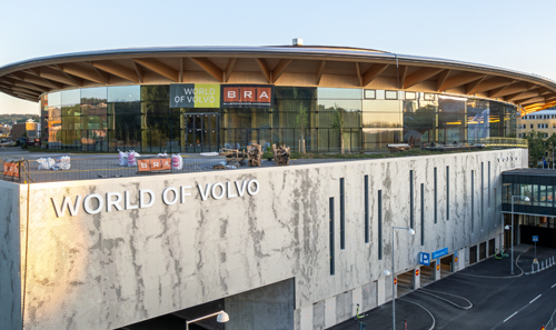 A modern hotel room with two neatly made twin beds, a wall-mounted TV, a desk with a chair, and a large window overlooking a city at night. The room has wooden floors, gray and white decor, and soft lighting from a bedside lamp.