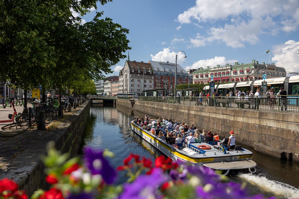 A modern hotel room with two neatly made twin beds, a wall-mounted TV, a desk with a chair, and a large window overlooking a city at night. The room has wooden floors, gray and white decor, and soft lighting from a bedside lamp.