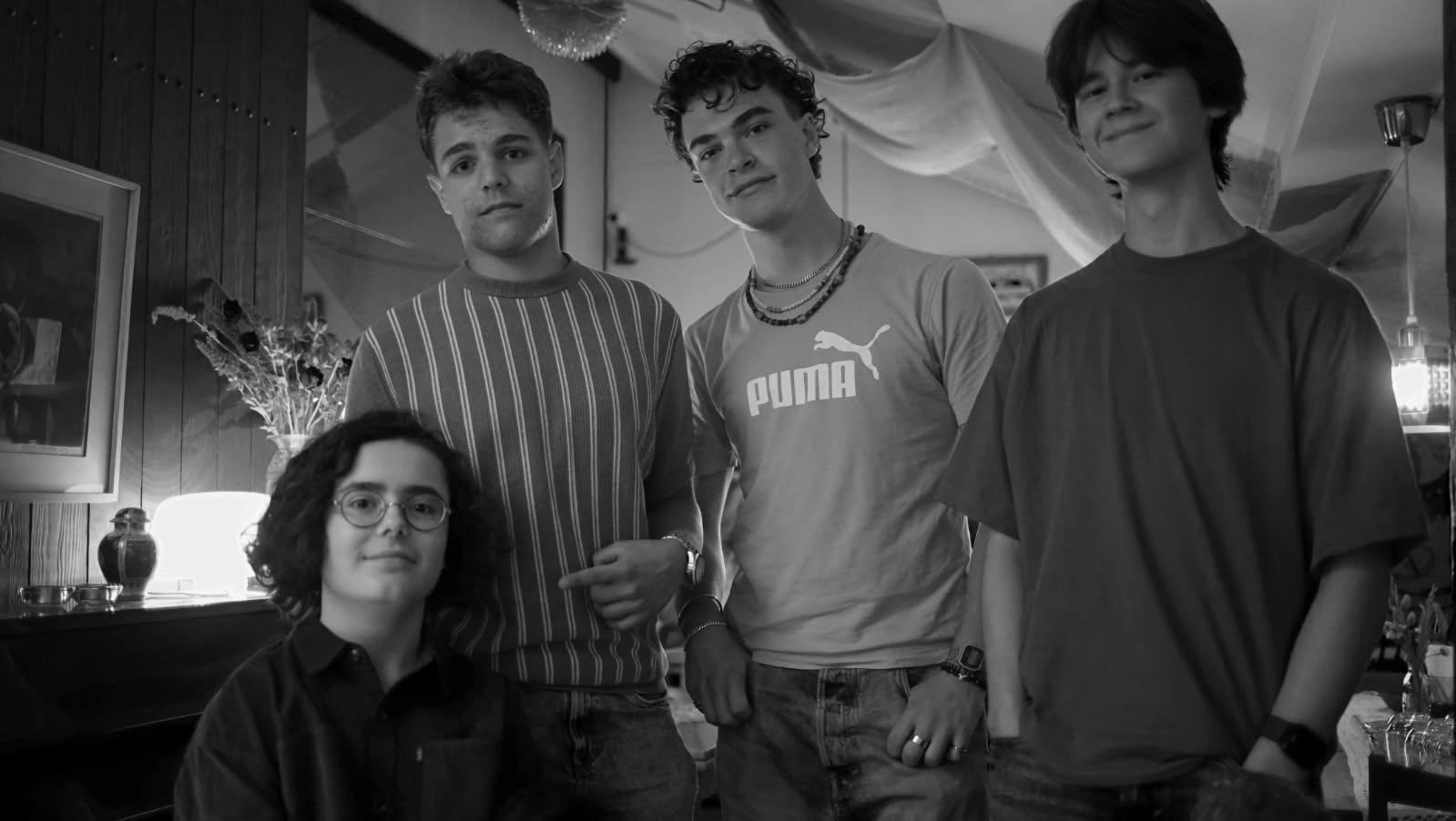 A modern hotel room with two neatly made twin beds, a wall-mounted TV, a desk with a chair, and a large window overlooking a city at night. The room has wooden floors, gray and white decor, and soft lighting from a bedside lamp.