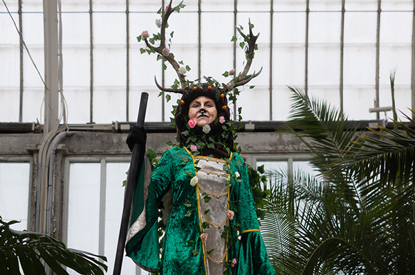 A modern hotel room with two neatly made twin beds, a wall-mounted TV, a desk with a chair, and a large window overlooking a city at night. The room has wooden floors, gray and white decor, and soft lighting from a bedside lamp.