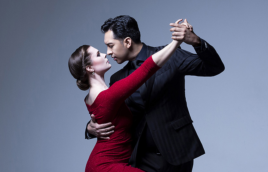 A modern hotel room with two neatly made twin beds, a wall-mounted TV, a desk with a chair, and a large window overlooking a city at night. The room has wooden floors, gray and white decor, and soft lighting from a bedside lamp.