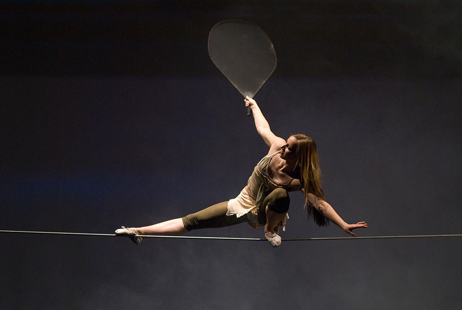 A modern hotel room with two neatly made twin beds, a wall-mounted TV, a desk with a chair, and a large window overlooking a city at night. The room has wooden floors, gray and white decor, and soft lighting from a bedside lamp.